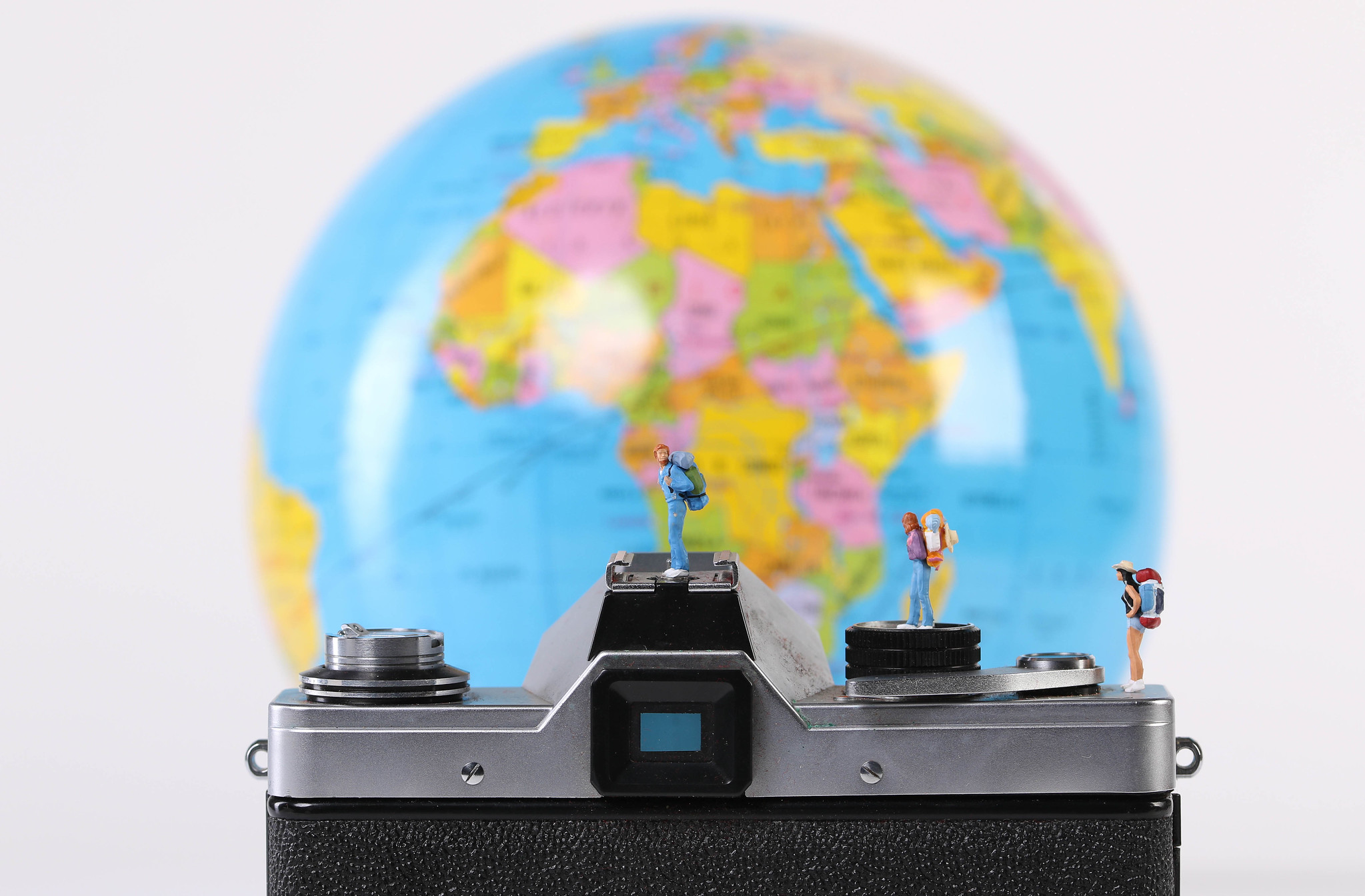 From the back of an old film camera, 4 tiny figures with backbacks look out at a globe in the backgound, slightly blurred from shallow depth of field, centered on Africa